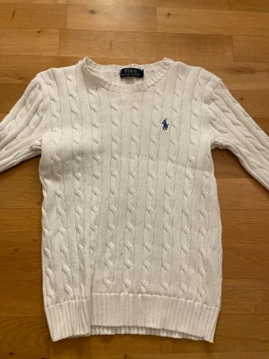 Vit kabelstickad Ralph Lauren - Kabelstickad vit tröja från Polo Ralph Lauren med rund halsringning och blå broderad logga på bröstet. Tröjan har långa ärmar och ribbade muddar vid ärmslut och nederkant. Klassisk och stilren design i mjukt material. Pris kan diskuteras!