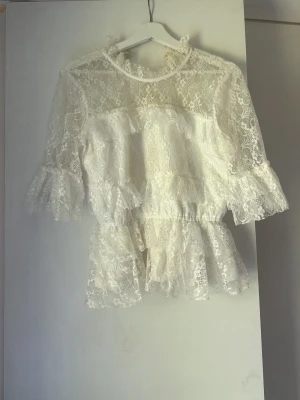 Vit spetsblus med volanger - Romantisk vit blus i genombruten spets med volangdetaljer vid ärmslut och midja. Blusen har hög halsringning och trekvartsärm, perfekt för en trendig och feminin look. Materialet är lätt och luftigt, vilket ger en elegant känsla. Liknar by Malians. 