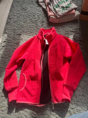Rosa fleecejacka från Polarn O. Pyret - Snygg röd fleecejacka från Polarn O. Pyret med dragkedja framtill och höga fickor på sidorna. Jackan har en hög krage och är perfekt för lager-på-lager under kyliga dagar. Tillverkad i mjuk fleece som känns skön mot huden. Perfekt för barn i 7-8 års åldern.