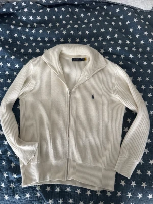 Vit ribbstickad kofta från Polo Ralph Lauren - Vit ribbstickad kofta med dragkedja från Polo Ralph Lauren. Klassisk krage och broderad logga på bröstet. Mjuk och skön, perfekt för lager-på-lager. Långärmad och i ett stilrent snitt som funkar till det mesta. Passar M och L