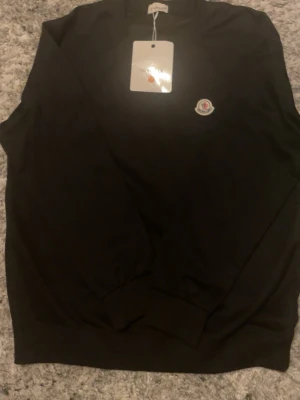 Svart stickad tröja från Moncler - Svart stickad tröja från Moncler med rund halsringning och diskret Moncler-logga på bröstet. Tröjan har långa ärmar och ribbade muddar vid ärmslut och nederkant. Perfekt för en stilren och enkel look.