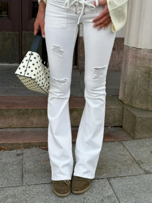 Vita bootcut jeans med snörning - Snygga vita bootcut jeans med snörning i midjan och slitna detaljer på låren. Jeansen har hög midja och utsvängda ben, vilket ger en trendig siluett. Perfekta för dig som vill sticka ut med en cool och modern look.