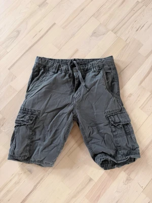 Grå cargoshorts med snörning - Säljer ett par grå cargoshorts med flera praktiska fickor på sidorna och baktill. Shortsen har snörning i midjan och är tillverkade i bomull, vilket gör dem sköna och lätta att bära. Perfekta för en avslappnad stil under varmare dagar.