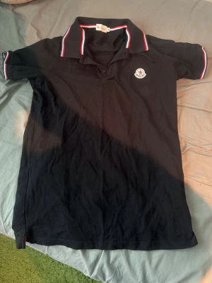 Moncler pikétröja M - Svart pikétröja Moncler med klassisk krage och knappar framtill. Tröjan har röda och vita ränder på krage och ärmslut samt Moncler-logga på bröstet. Perfekt för en stilren och sportig look.