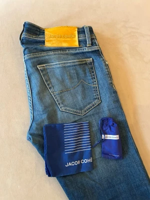 Blå jeans från Jacob Cohën - Snygga blå jeans från Jacob Cohën Nick med klassisk femficksdesign och kontrastsömmar i storlek 29. Jeansen har en gul patch med logga bak och levereras med tillbehör i blått tyg. Perfekta för en trendig och avslappnad stil.