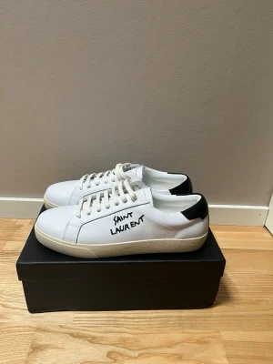 Vita Saint Laurent sneakers - Snygga vita sneakers från Saint Laurent med svart detalj på hälen och handskriven logga på sidan. Pris går att diskuteras, skriv vid funderingar! Dustbag och kort ingår