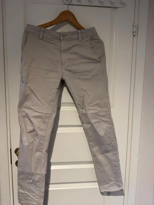 Grå/vita chinos - Snygga grå/vita chinos med klassisk passform och raka ben. Byxorna har framfickor, bälteshällor och två bakfickor med knapp. Perfekta för en clean och stilren look. Materialet är mjukt och bekvämt, troligtvis bomull.