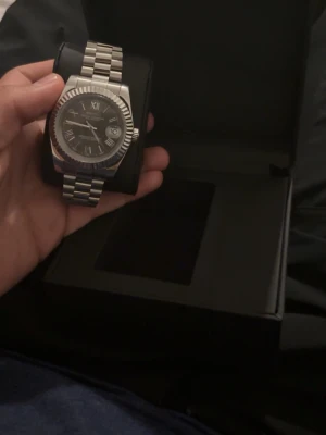 Seiko klocka - Stilren Seiko klocka i silverfärgat stål med klassisk länkarmband och svart urtavla. Klockan har romerska siffror, datumvisning och en markerad räfflad bezel. Perfekt accessoar för dig som gillar en tidlös och elegant look. Modell daydate ny pris 4000.