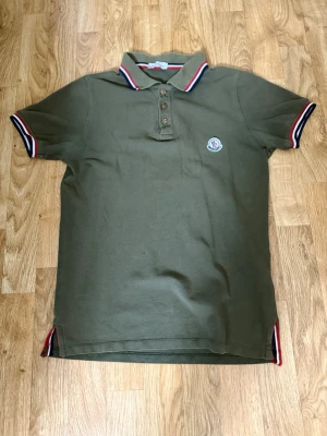 Olivgrön Moncler pikétröja - Pris kan diskuteras vid snabb affär .Snygg olivgrön pikétröja från Moncler med klassisk krage och knappar framtill. Trikolorfärgade detaljer på krage, ärmslut och sidor. Liten Moncler-logga på bröstet. Perfekt för en stilren och sportig look.