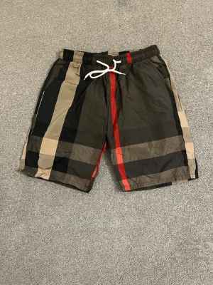 Rutig shorts med snörning från Burberry - Snygga shorts från Burberry med klassiskt rutigt mönster i beige, svart och rött. De har elastisk midja med vit snörning och är tillverkade i ett lätt syntetmaterial. Perfekta för sommaren och ger en trendig look.