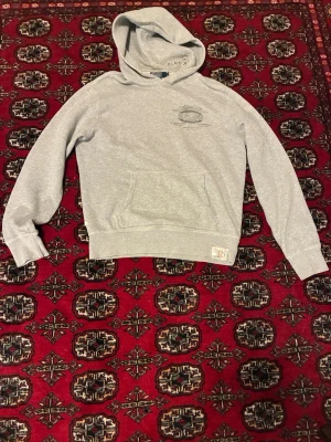 Polo Ralph Lauren hoodie vintage  - Snygg ljusgrå hoodie från polo med klassisk huva och magficka. Diskret logga på bröstet och ribbade muddar vid ärmslut och nederkant. Tillverkad i mjuk bomullsblandning som känns skön mot huden. Perfekt för en avslappnad och stilren look.
