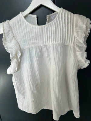 Vit blus med volangärmar - Superfin vit blus med volangdetaljer på ärmarna och plisserad överdel. Blusen har rund halsringning och är tillverkad i ett lätt, luftigt tyg med struktur. Perfekt för en stilren och trendig look.