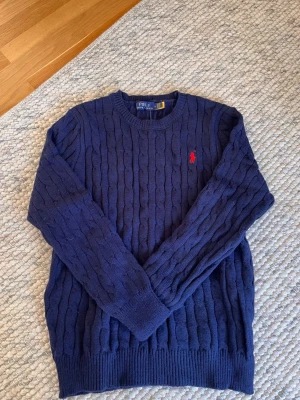 Marinblå kabelstickad tröja Polo Ralph Lauren - Snygg marinblå kabelstickad tröja från Polo Ralph Lauren med klassisk röd logga broderad på bröstet. Tröjan har rund halsringning, ribbade muddar och är tillverkad i mjukt material. Perfekt för en stilren och avslappnad look.