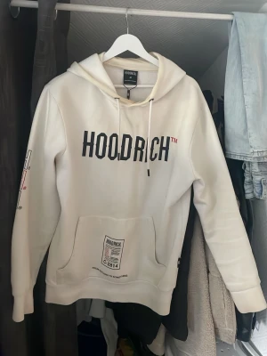 Vit hoodie från Hoodrich med tryck - Snygg vit hoodie från Hoodrich med svart logotyptryck på bröstet och detaljer på fickan och ärmen. Klassisk känguruficka framtill och justerbar huva med dragsko. Perfekt för en streetwear-look.