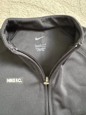 Svart Nike F.C. ziptröja - Snygg svart ziptröja från Nike F.C. med hög krage och hel dragkedja framtill. Tröjan har diskret Nike-logga på bröstet och två fickor. Tillverkad i mjukt och stretchigt material, perfekt för träning eller chill.  Finns 2 st