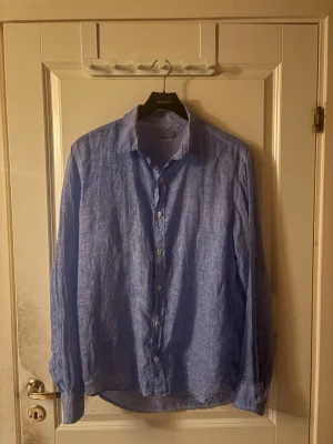 Ljusblå linneskjorta  - Snygg ljusblå skjorta i 100%. Skjortan har klassisk krage, långa ärmar med manschett och knäppning framtill. Perfekt för en avslappnad men stilren look. 