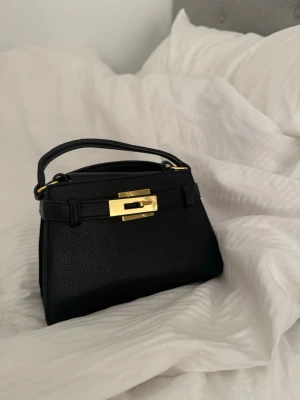 Svart minihandväska med gulddetaljer - Stilren svart minihandväska i läderimitation med elegant guldfärgat spänne och handtag upptill. Väskan har en klassisk, boxig form och passar perfekt för dig som gillar minimalistisk och trendig design.