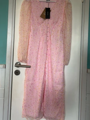 Rosa blommig långklänning vero moda - Söt rosa långklänning från vero moda med små blommor i gult och lila. Klänningen har långa, transparenta ärmar och v-ringning. Tillverkad i ett lätt och luftigt polyestertyg, perfekt för varmare dagar. Nypris 499