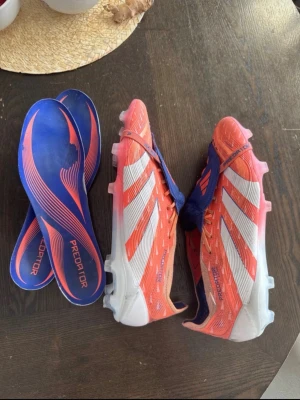 Adidas Predator fotbollsskor elite tounge FG/AG - PRIS KAN DISKUTERAS! Säljer ett par Adidas Predator fotbollsskor i rött, vitt och blått. Skorna har klassisk design med vita ränder, blå detaljer och utbytbara innersulor. Yttermaterialet är syntet och sulan har fasta dobbar för spel på gräs och konstgräs. Jag säljer dessa enbart för att de passade snäppet tajt och därför rekommenderar jag att om man har 45,5/45 kommer dessa sitta perfekt men också 46 ifall man gillar den passformen