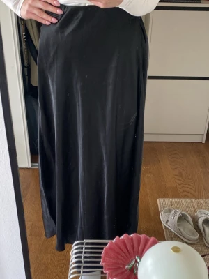 Svart långkjol i satin - Stilren svart långkjol i glansigt satinliknande material. Kjolen har en rak och enkel passform som faller snyggt längs kroppen. Aldrig använd.