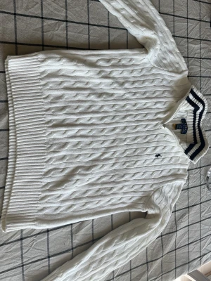 Vit stickad tröja Polo Ralph Lauren - Säljer en vit stickad tröja från Polo Ralph Lauren med klassiskt kabelmönster och halv dragkedja vid halsen. Tröjan har ribbade muddar och en broderad logga på bröstet. Perfekt för dig som gillar stilrena och tidlösa plagg. Nypris 1600, tröjan är använd max 2-3 gånger 