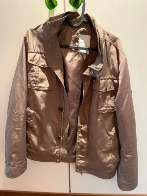 Beige vindjacka med fickor - Stilren beige vindjacka med ståkrage och flera praktiska fickor framtill. Jackan har knappar och dragkedja samt justerbara detaljer i midjan. Perfekt för dig som gillar en enkel och clean look.