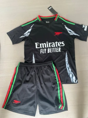 Arsenal svart träningsset - Snyggt svart träningsset från Arsenal med röd och grön detalj på krage och shorts. Setet består av t-shirt och shorts i lätt och snabbtorkande material. Arsenal-logga i rött och Emirates Fly Better-tryck på tröjan. Perfekt för fotboll eller gym.