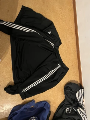 Svart Adidas sweatshirt 2XL - Säljer en svart sweatshirt från Adidas i storlek 2XL. Tröjan har de klassiska vita ränderna längs ärmarna och Adidas-loggan på bröstet. Tillverkad i ett mjukt och bekvämt material, perfekt för träning eller chill. Enkel och stilren design som funkar till det mesta.