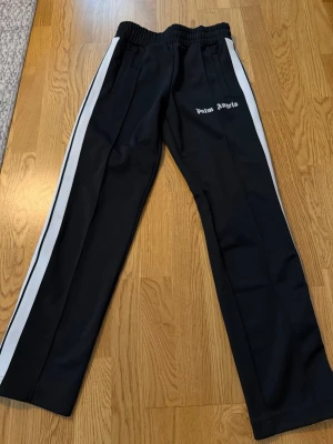 Palm Angels svarta mjukisbyxor - Svarta mjukisbyxor från Palm Angels med vita sidoränder och elastisk midja. Byxorna har en avslappnad passform och loggan broderad framtill. Perfekta för en streetwear-look och tillverkade i ett mjukt, sportigt material.