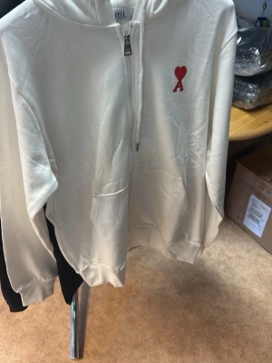 Vit hoodie med röd Ami-dekal - Snygg vit hoodie från Ami med dragkedja framtill och klassisk huva. På bröstet sitter en röd Ami-logga med hjärta. Hoodien har fickor på sidorna och ribbade muddar. Perfekt för en clean och stilren look.