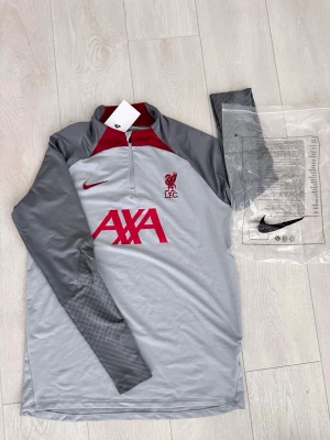 Grå Liverpool träningströja Nike - Snygg långärmad träningströja från Nike i grått med röda detaljer, Liverpool FC-logga och AXA-tryck på bröstet. Halv dragkedja framtill och kontrasterande mörkgrå ärmar. Perfekt för fotbollsträning eller gymmet.