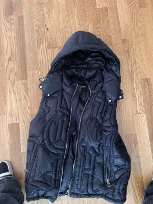 Demont väst - Snygg svart dunväst från demont med avtagbar huva och coola quiltade detaljer. Västen har två dragkedjefickor framtill och stängs med dragkedja. Perfekt för lager-på-lager och streetstyle. Materialet är glansigt och ger en modern look.