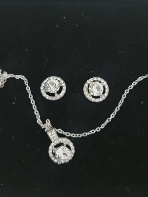 Smyckeset med gnistrande stenar - Ett stilrent smyckeset med halsband och örhängen i silverfärg. Både halsbandet och örhängena har runda hängen med klara gnistrande stenar omgivna av mindre stenar. Perfekt för dig som gillar bling och eleganta accessoarer.