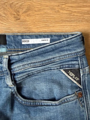 Blå jeans Replay Rocco Comfort Fit - Säljer ett par klassiska blå jeans från Replay, modell Rocco Comfort Fit. Jeansen har normal midja, raka ben och snygga kontrastsömmar. Tillverkade i slitstark denim med detaljer som Replay-logga på fickan och metallknapp framtill.