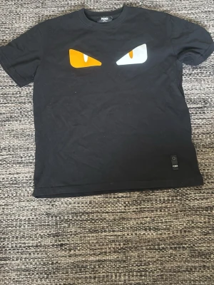 Svart Fendi t-shirt med gul logga - Snygg svart t-shirt från Fendi med gul logga tryckt i nacken och en liten Fendi-detalj med knapp längst ner. Klassisk passform och rund halsringning, perfekt för dig som gillar stilrena designer med exklusiv känsla.