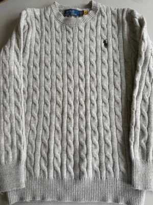 Ljusgrå kabelstickad tröja Polo Ralph Lauren - Kabelstickad ljusgrå tröja från Polo Ralph Lauren med rund halsringning och broderad logga på bröstet. Tröjan har ribbade muddar vid ärmslut och nederkant. Perfekt för lager-på-lager och klassisk stil.