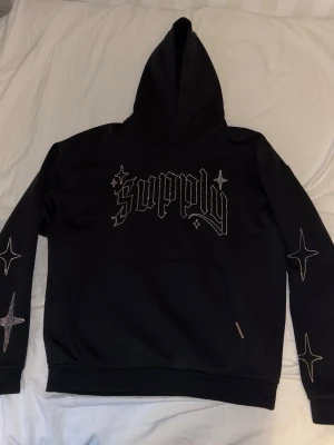 Svart hoodie Supply med stjärnor - Svart hoodie från Supply med stor huva och magficka. Framsidan har ett stort tryck med texten 'supply' i gotisk stil och flera stjärndetaljer i vitt på både bröstet och ärmarna. Tillverkad i mjukt material, perfekt för en avslappnad streetwear-look.