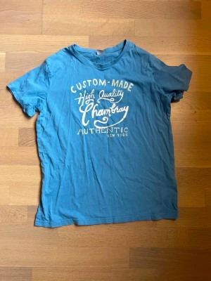 Blå t-shirt med tryck Chambray - Blå t-shirt från Chambray med vitt tryck framtill där det står 'Custom-Made High Quality Chambray Authentic New York'. Klassisk passform och rund halsringning, perfekt för en avslappnad stil.