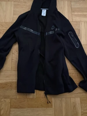 Svart hoodie från Nike tech med dragkedja - Svart hoodie från Nike med hel dragkedja framtill och huva. Snygga detaljer med svarta tejpade dragkedjor på bröstet och ärmen. Tillverkad i mjukt material och har en sportig look. Perfekt för dig som gillar streetwear och vill ha något stilrent.