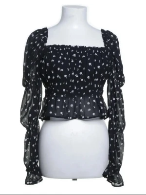 Svart blommig offshoulderblus - Trendig svart offshoulderblus från H&M med små vita blommor. Blusen har långa puffärmar med resår vid ärmslut och midja, samt volangdetaljer längs halsringningen. Tillverkad i ett tunt, transparent material som ger en luftig känsla.