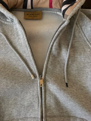 Grå zip hoodie från Burberry - Snygg grå zip hoodie från Burberry med klassiskt rutigt foder i huvan och dragsko. Tillverkad i mjukt bomullsmaterial och har en rejäl dragkedja med Burberry-logga. Perfekt för dig som gillar stilrena och exklusiva plagg.