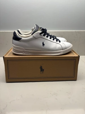 Vita Polo Ralph Lauren sneakers(låda ingår ej) - Stilrena vita sneakers från Polo Ralph Lauren med mörkblå detaljer och logga på sidan. Skorna har mörkblå snören, vit sula och är tillverkade i slätt läder. Klassisk låg modell som passar till det mesta och ger en clean look.