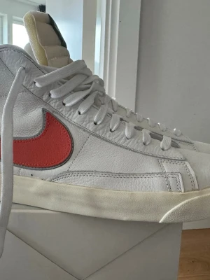 Nike Blazer Mid 77 Vintage Vit/Röd - Nike Blazer Mid 77 Vintage sneakers i vitt läder med röd Swoosh och retro-vibe. Skorna har högt skaft, vit sula och gröna detaljer på plösen. Klassisk siluett med robust snörning och tidlös design.