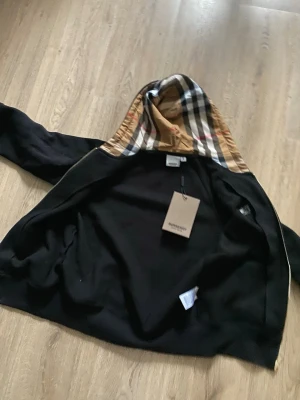 Svart hoodie från Burberry med rutig huva - Snygg svart hoodie från Burberry med den klassiska beige, vita och röda rutan i huvan. Dragkedja framtill och mjukt material som känns lyxigt. Perfekt för dig som vill ha en exklusiv streetwear-look med ikonisk design och detaljer.