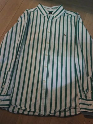 Randig skjorta från Polo Ralph Lauren - Snygg vit skjorta med gröna vertikala ränder från Polo Ralph Lauren. Klassisk krage, långärmad och med knappar framtill. Liten broderad logga på bröstet. Tillverkad i bomull med naturlig stretch för extra komfort.