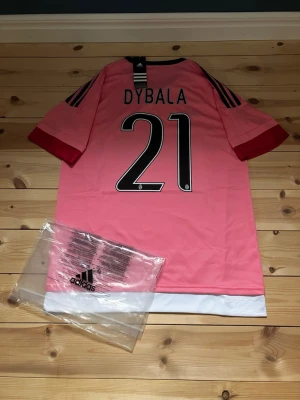 Juventus rosa matchtröja Dybala #21 - Juventus rosa matchtröja från Adidas med Dybala och nummer 21 på ryggen. Tröjan har svarta detaljer, vita och röda inslag samt Jeep-logga på bröstet. Tillverkad i lätt och ventilerande climacool-material, perfekt för fotboll eller träning.