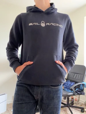 Marinblå hoodie från Sail Racing - Snygg marinblå hoodie från Sail Racing med vit logga tryckt på bröstet. Klassisk känguruficka framtill och justerbar huva med dragsko. Mjuk insida i fleece för extra komfort. Perfekt för chill dagar eller när du vill ha en sportig look.