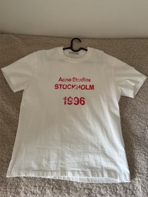 Acne Studios t-shirt - Vit t-shirt från Acne Studios med rött tryck på bröstet där det står 'Acne Studios STOCKHOLM 1996'. Klassisk passform och rund halsringning. Tillverkad i mjuk bomull, perfekt för en clean och stilren look.