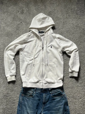 Vit hoodie från Polo Ralph Lauren - Säljer en stilren vit hoodie från Polo Ralph Lauren med dragkedja, två fickor fram och klassisk marinblå logga på bröstet. Hoodien har snörning i huvan och ribbade muddar vid ärmar och nederkant. Perfekt för en avslappnad look. Skulle påstå storlek small!