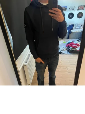 Svart hoodie från Calvin Klein - Säljer en svart hoodie från Calvin Klein med dragsko i huvan och CK-logga på vänster ärm. Klassisk passform och mjukt material som känns skönt mot huden. Perfekt för dig som gillar stilrena och enkla plagg med en touch av märke.